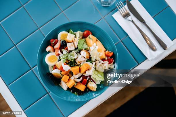 flat lay of a serving of healthy gourmet fresh and colourful salad bowl with organic greens, pumpkin, cherry tomatoes, cucumber, boiled egg, grilled chicken, raspberry and strawberry on blue tile table. healthy eating, go green lifestyle - china balance stock-fotos und bilder