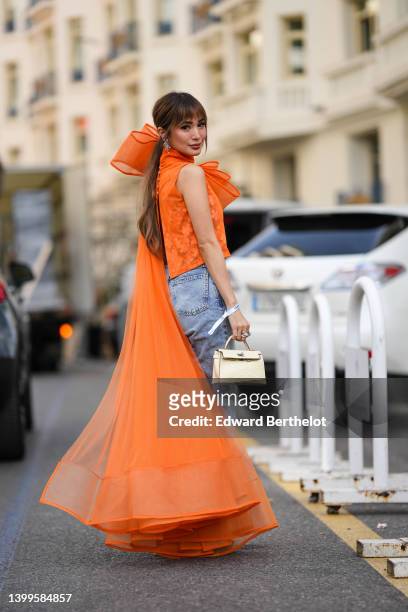 Heart Evangelista wears jewelry, earrings, an orange turtleneck top with floral pattern embroidery, a tulle cape, blue denim jeans, a Hermes bag,...