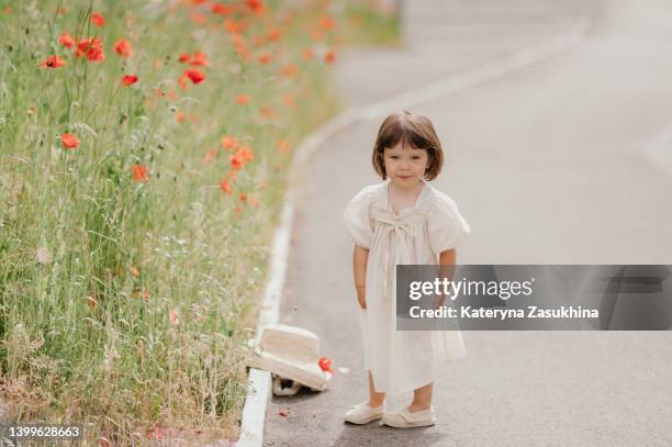 a photo of a little baby girl in a summer dress walking in the street near poppy flowers on a sunny day - toddler girl dress stockfoto's en -beelden