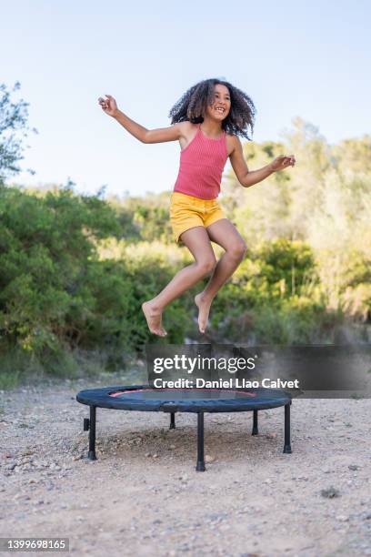 young girl with curly hair having fun while jumping on a trampoline outdoors in nature. - trampolin stock-fotos und bilder