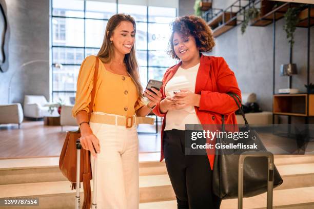 two smiling businesswomen using smart phones to share files in a hotel lobby - partilha de ficheiros imagens e fotografias de stock