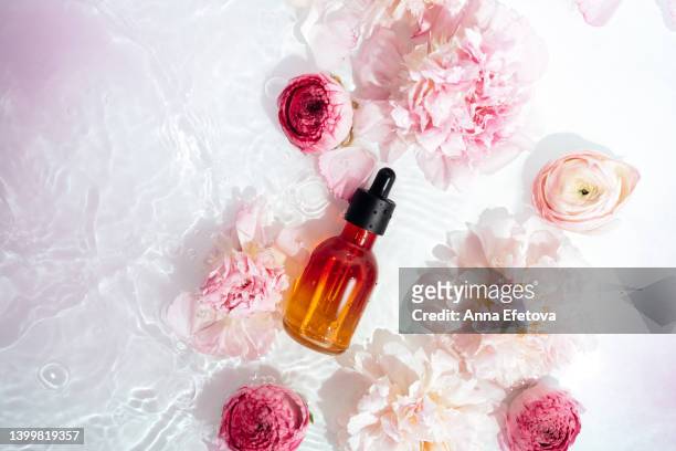 orange glass bottle of face serum with polyglutamic acid is lying in water on white background with gentle pink peonies and ranunculus. beautiful backdrop for your design with copy space. macro photography from above - white rose flower spa photos et images de collection