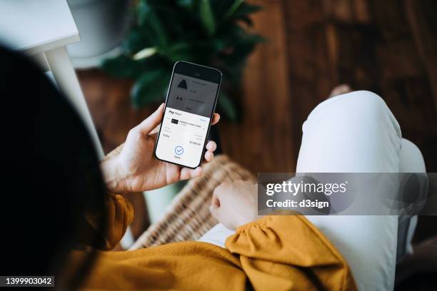 over the shoulder view of young asian woman relaxing at home, sitting on the armchair in the living room, shopping online with smartphone and making payment online with credit card. lifestyle and technology. comfortable and fast shopping experience - shopping screen stockfoto's en -beelden