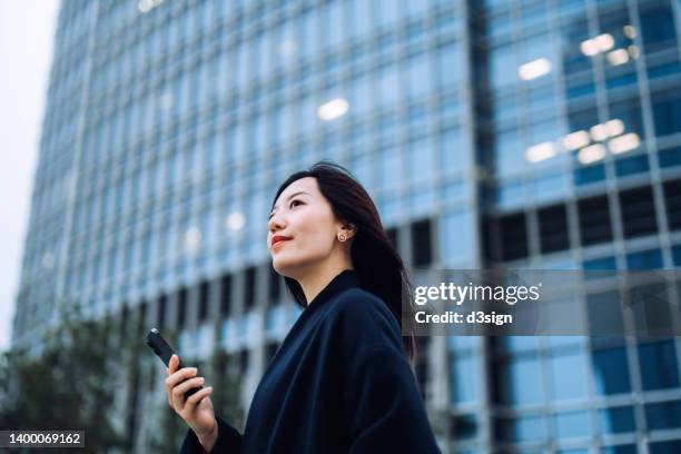 confident and determined young asian businesswoman looking ahead with smile, holding smartphone while commuting in downtown district against contemporary corporate skyscrapers in city. business lifestyle with technology, staying connected anytime anywhere - business smile stockfoto's en -beelden