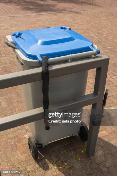 gray waste bin with two black wheels secured with a padlock to a steel fence in the street - prullenbak op wielen stockfoto's en -beelden