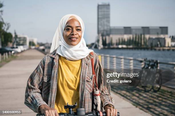 young smiling woman with hijab on e-scooter at river promenade in modern berlin - islamic action front stock pictures, royalty-free photos & images