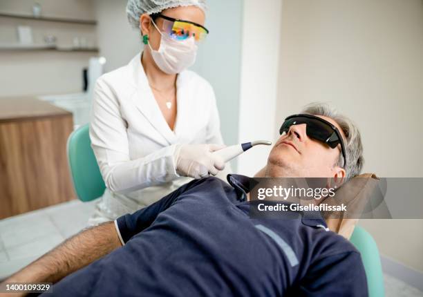 homem adulto médio recebendo tratamento a laser por um cosmetólogo em clínica - máscara cirúrgica - fotografias e filmes do acervo