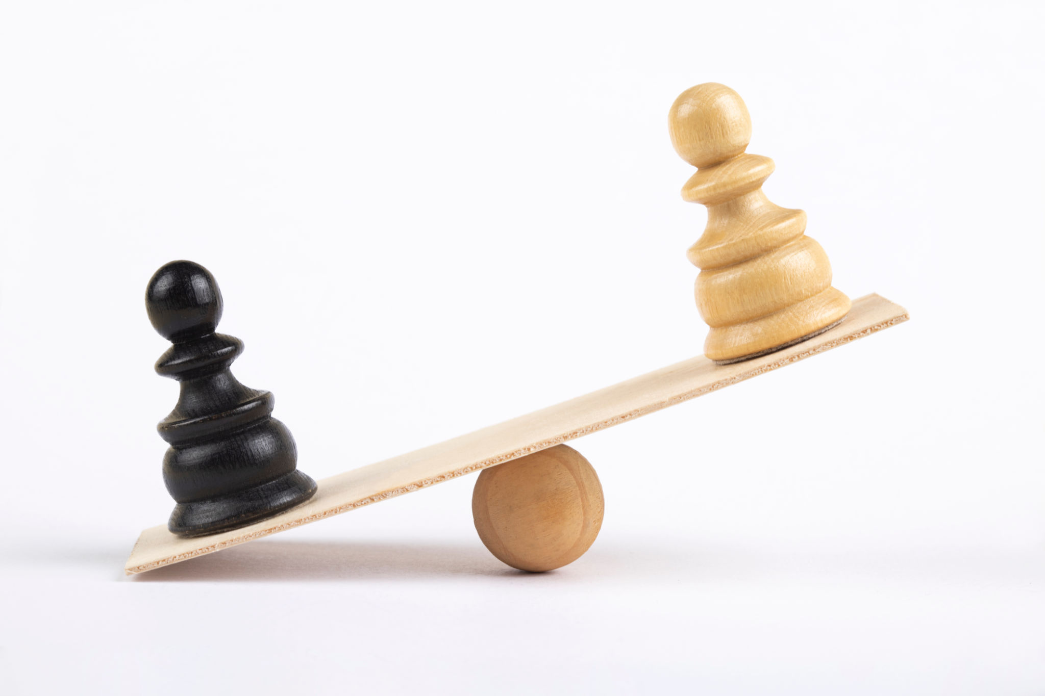 White and a black pawn chess piece stand on wooden seesaw White and a black pawn chess piece stand on wooden seesaw