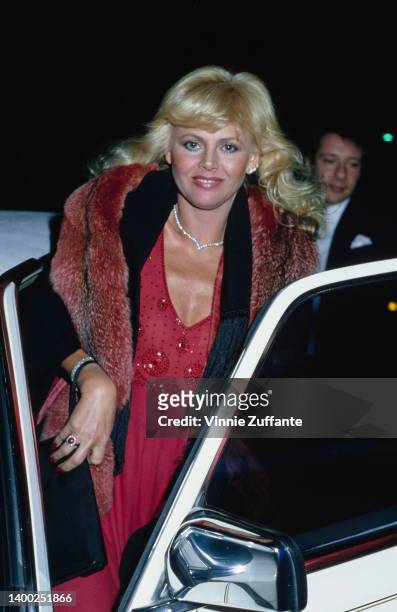 Swedish actress and singer Britt Ekland, holding a red fur coat over a red dress as she gets in an car, United States, December 1981.