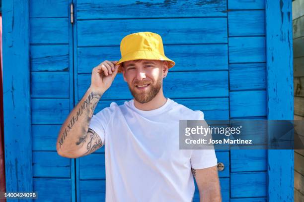 man on vacation with a blue colored beach hut behind him - bucket hat stock pictures, royalty-free photos & images