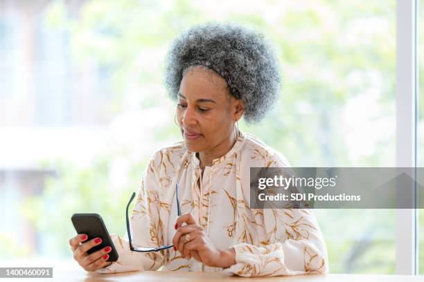 senior adult woman reads a message on her phone - black woman grey hair stock pictures, royalty-free photos & images