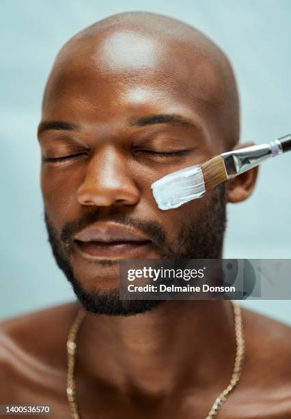 african american male model applying lotion with a brush on his face. the man has a beard and bald head. he is shirtless and wearing a necklace - man eye cream imagens e fotografias de stock