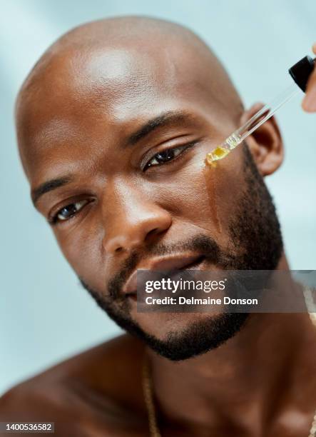 afrikanischer mann, der mit einer ölpipette serum auf sein gesicht aufträgt. er macht seine morgendliche hautpflegeroutine. das schönheitsmodell hat eine klare und saubere haut - african mask stock-fotos und bilder