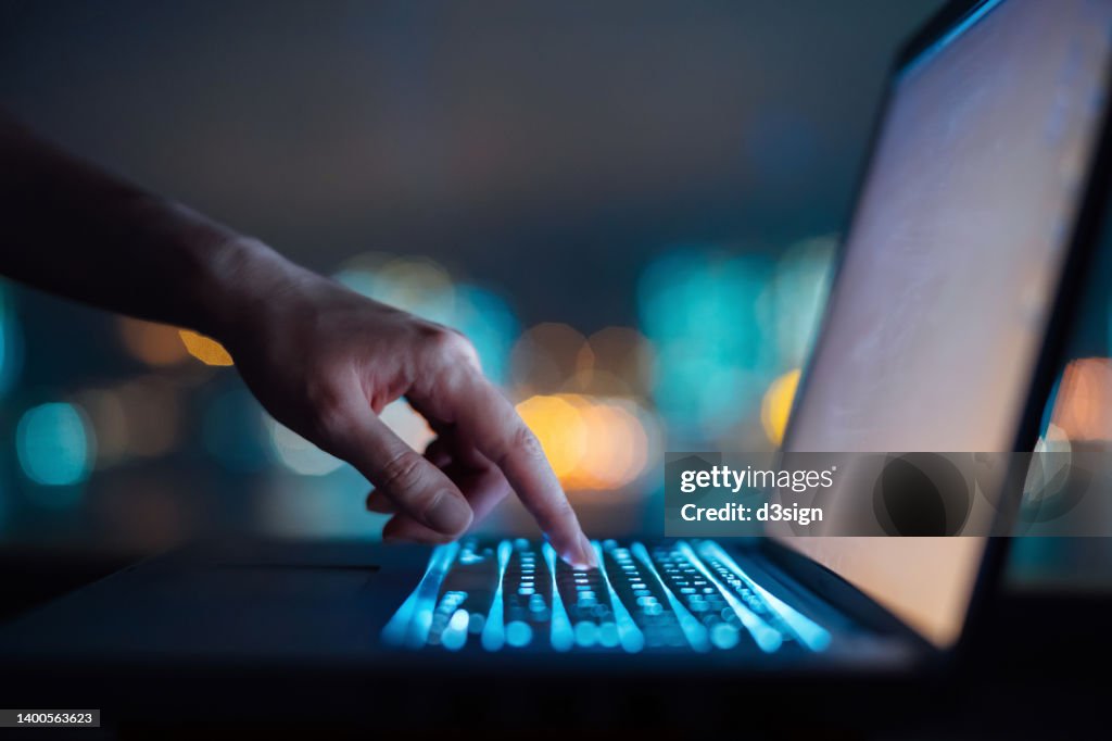 Close up of woman's hand typing on computer keyboard in the dark against colourful bokeh in background, working late on laptop at home