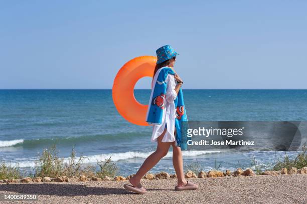 woman walking towards the beach with an orange float and summer clothes - toalla stock pictures, royalty-free photos & images