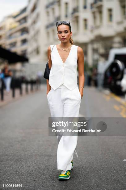 Hana Cross wears sunglasses, a sleeveless white jacket, earrings a golden necklace, a black bag, white flare pants, green and yellow sneakers shoes,...