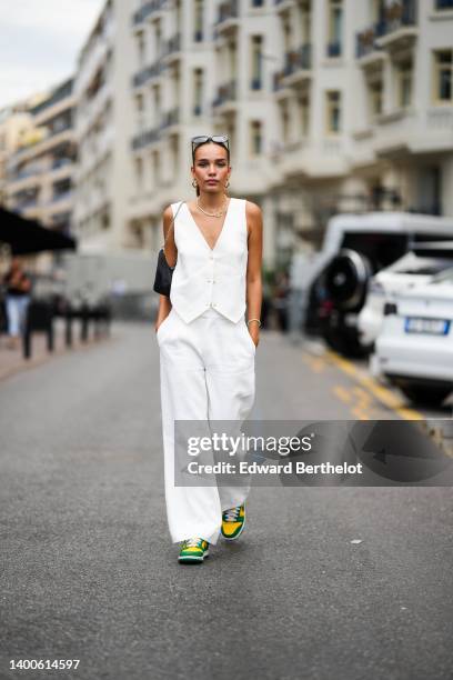 Hana Cross wears sunglasses, a sleeveless white jacket, earrings a golden necklace, a black bag, white flare pants, green and yellow sneakers shoes,...