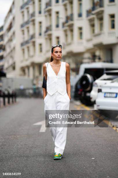 Hana Cross wears sunglasses, a sleeveless white jacket, earrings a golden necklace, a black bag, white flare pants, green and yellow sneakers shoes,...