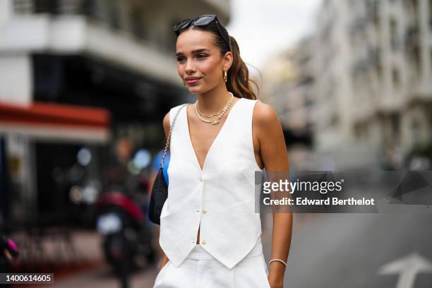 Hana Cross wears sunglasses, a sleeveless white jacket, earrings a golden necklace, a black bag, white flare pants, on May 26, 2022 in Cannes, France.