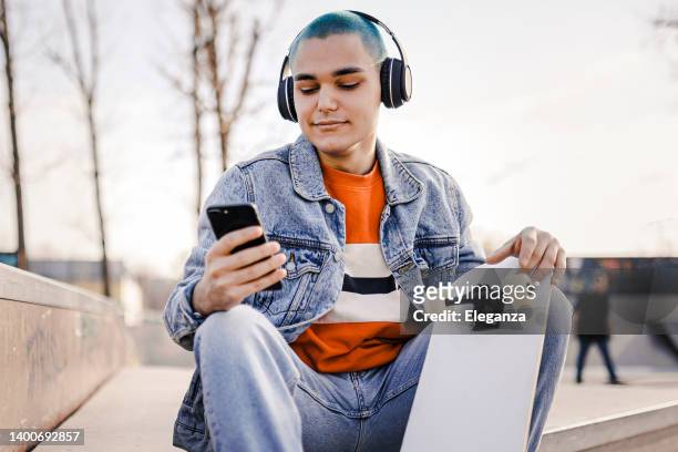 boy  relaxing with his skateboard listening to music. urban boy enjoying listening to music from her mobile phone while sitting at skatepark. - young man listening to music on smart phone outdoors stockfoto's en -beelden