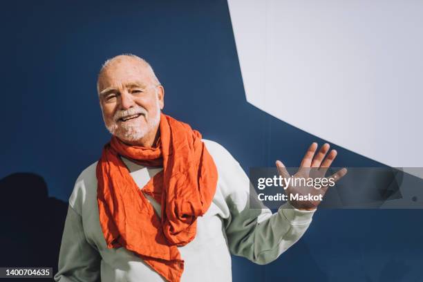 portrait of happy senior man wearing scarf waving hand against blue and white wall - zwaaien stockfoto's en -beelden