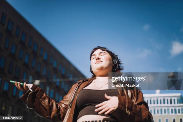 young woman with eyes closed listening music through wireless headphones during sunny day - body positivity stock-fotos und bilder