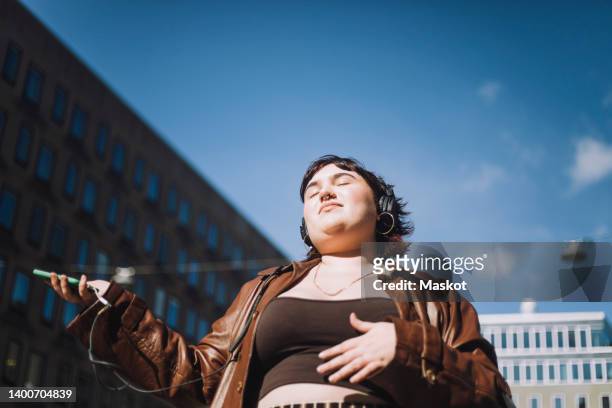 young woman with eyes closed listening music through wireless headphones during sunny day - body positive 個照片及圖片檔