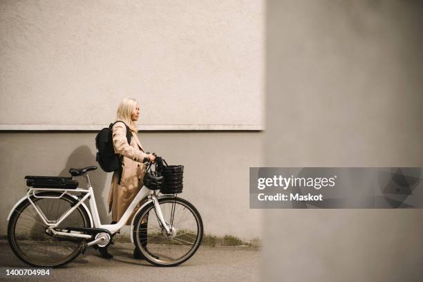 side view of businesswoman wheeling with electric bicycle by wall - ebike stock pictures, royalty-free photos & images