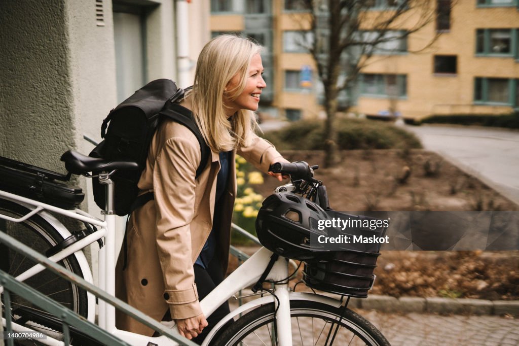 Side view of smiling businesswoman with backpack holding electric bicycle