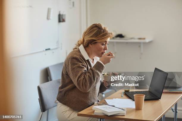 female professor eating lunch while sitting at desk with laptop in classroom - hungry stock pictures, royalty-free photos & images