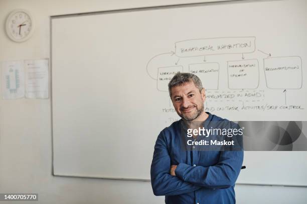 portrait of confident male teacher standing with arms crossed in front of whiteboard at university classroom - lecturer whiteboard stock pictures, royalty-free photos & images