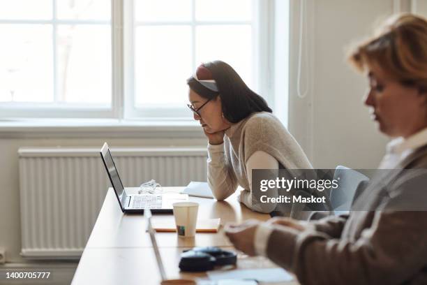 side view of female teacher watching laptop while sitting by colleague at desk in staffroom - leaning on elbows stock pictures, royalty-free photos & images
