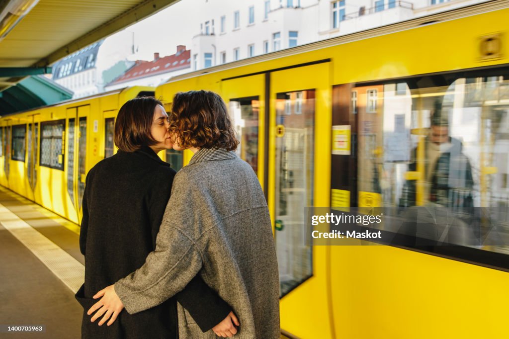 Lesbian couple kissing while standing by yellow train at railroad station
