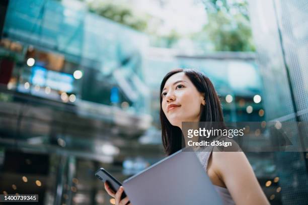 successful and modern young asian businesswoman carrying smartphone and laptop, commuting to work in central business district against contemporary corporate buildings in the city. female leadership. business on the go - erwartung stock-fotos und bilder