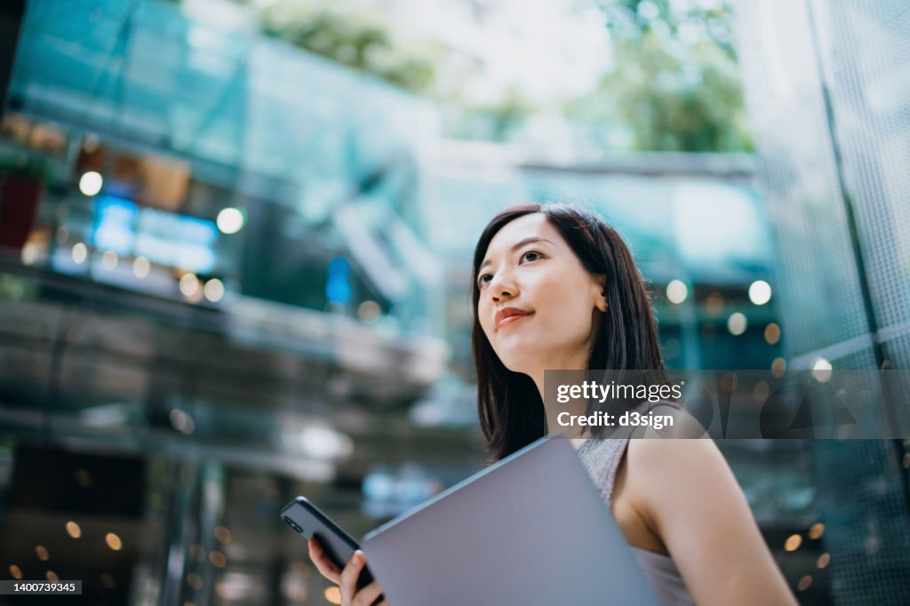 Successful and modern young Asian businesswoman carrying smartphone and laptop, commuting to work in central business district against contemporary corporate buildings in the city. Female leadership. Business on the go