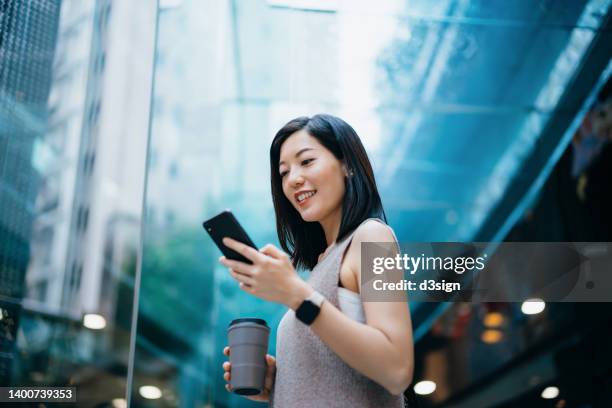 successful young asian businesswoman holding a reusable coffee cup, smiling after reading good news on her smartphone in contemporary office building. accomplishment and achievement at work. business on the go - will call stock pictures, royalty-free photos & images