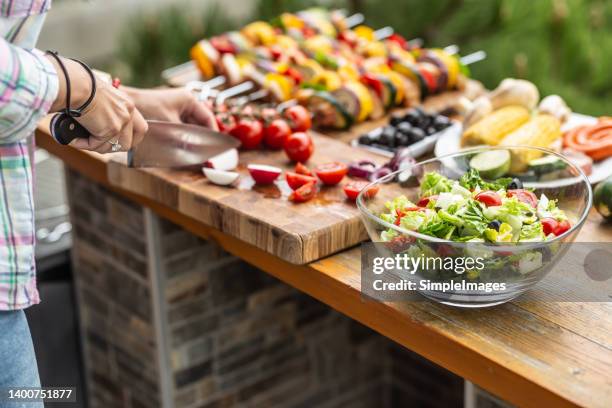 garden table full of food ready for a barbeque party with a salad in the front and a woman cutting vegetable aside. - barbeque party woman stock-fotos und bilder