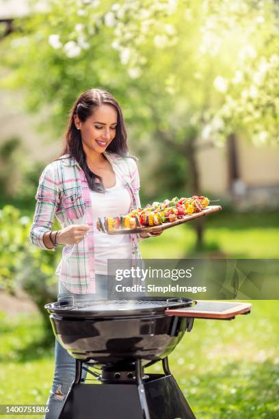 beautiful smiling woman brings meat and veg skewers to a hot smoking charcoal barbeque for preparation. - bbq chicken stock pictures, royalty-free photos & images