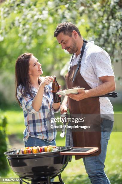 woman opens a bottle of beer to a man who holds tongs preparing food on a grill during a garden party. - couple grilling stock pictures, royalty-free photos & images