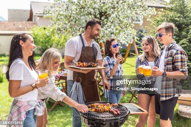 a group of friends with beers and drinks in their hands are having a good time chatting and standing around a charcoal barbeque. - barbeque party woman stock-fotos und bilder