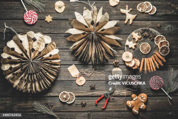 various christmas decoration  with handmade craft paper stars garlands, dried orange slices, lollipop, cookies, fir green and gingerbread man at rustic wooden table background - paper decoration stock-fotos und bilder