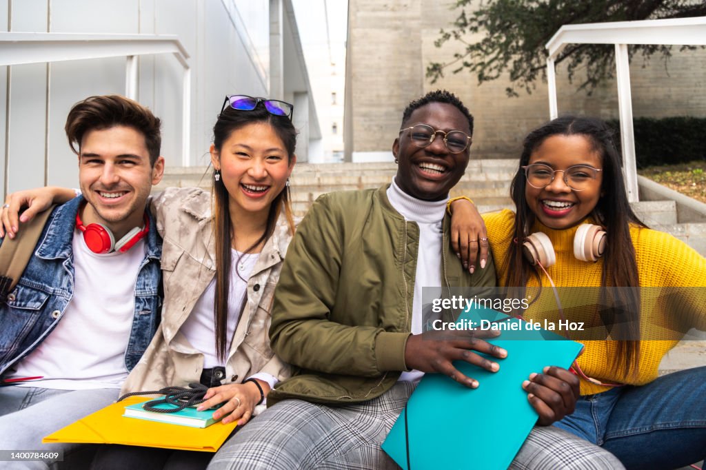 Happy and smiling group of multiracial college student friends looking at camera sitting on stairs outside university building.