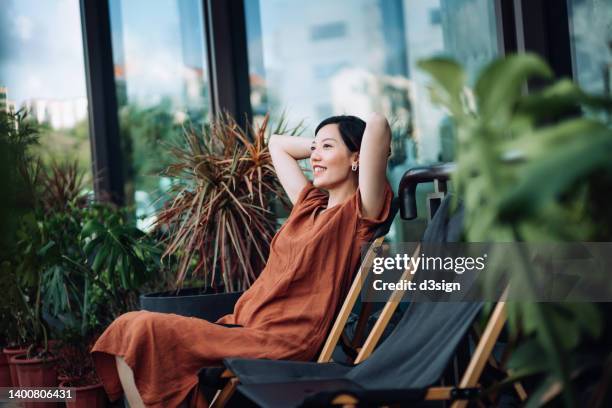 carefree young asian woman with her arms behind her head relaxing on deck chair in the balcony, enjoying summer day surrounded by beautiful houseplants - asian woman at home stock pictures, royalty-free photos & images