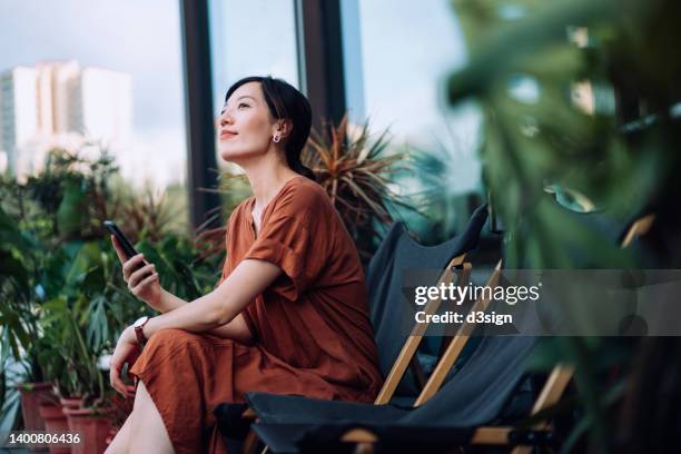 beautiful smiling young asian woman having a relaxing morning in the balcony of her apartment, sitting on deck chair and using smartphone, surrounded by beautiful houseplants. texting, leisure and lifestyle. technology in everyday life - booking hotel foto e immagini stock