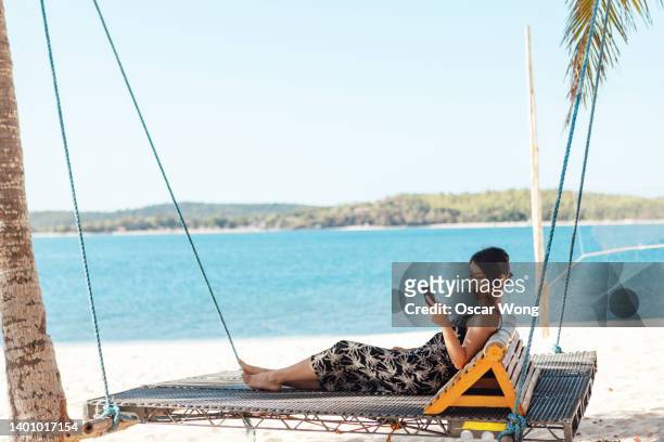 young asian woman using smart phone while relaxing on swing chair by the beach - smartphones dangling stock pictures, royalty-free photos & images