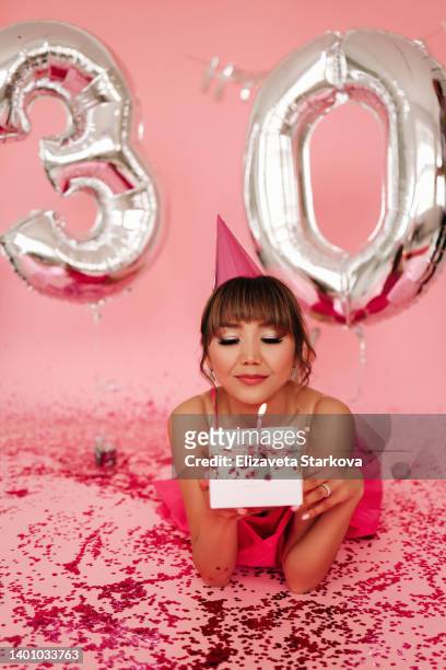 a beautiful happy joyful young woman in a bright pink mini dress is holding a cake laughing smiling celebrating her thirtieth birthday among confetti balloons on a pink background in a photo studio. a cheerful millennial girl in an elegant outfit - asian pin up girls stock-fotos und bilder