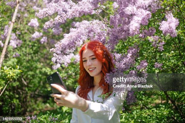 beautiful female portrait made outdoors in spring park in sunny day. young woman with long red curly hair making selfie with phone near lilac bush. - ginger bush stock pictures, royalty-free photos & images