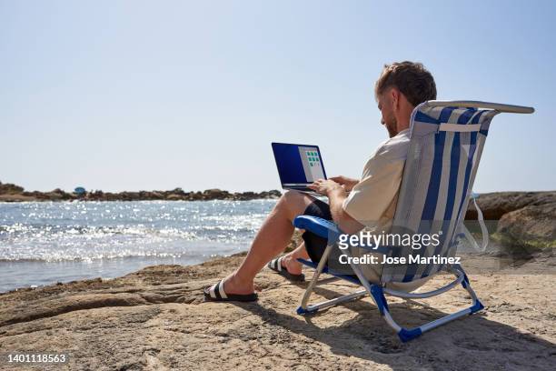 young man teleworking with his laptop from the beach sitting on a chair - abseits stock-fotos und bilder