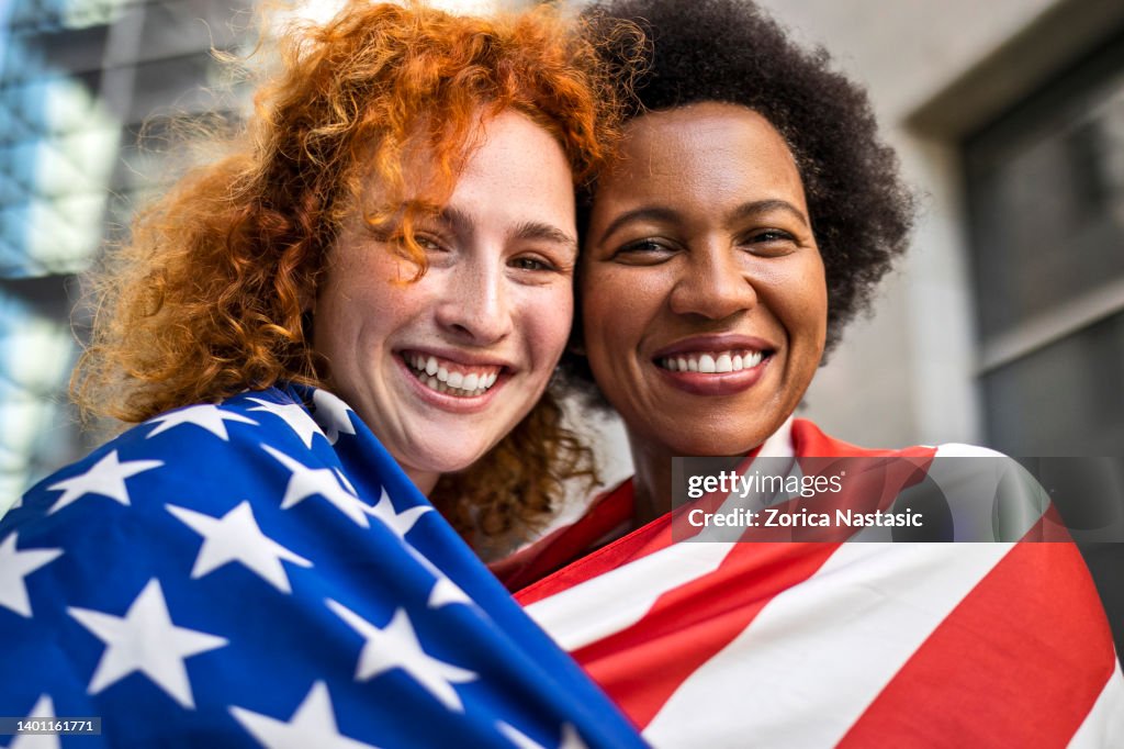 Girlfriends celebrating the 4th of July covered with American flag looking at camera