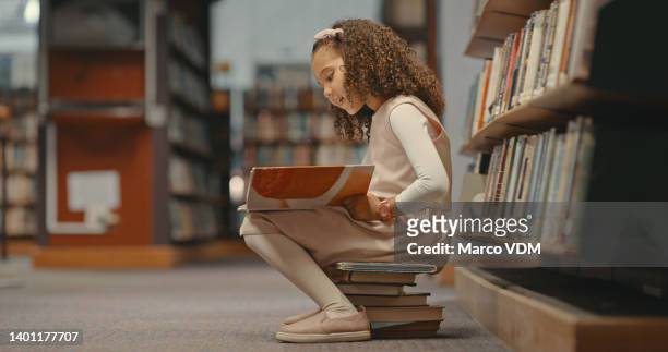 young girl sitting on books in the library and reading a book. cute girl with curly hair doing her project. female alone and doing research for a project - kids read stockfoto's en -beelden
